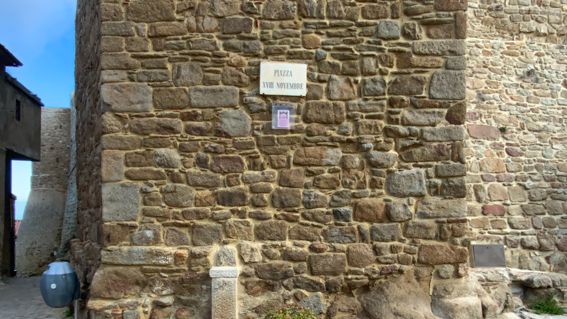 L'immagine rappresenta un muro in pietra con una targa bianca con scritto PIAZZA XVIII NOVEMBRE