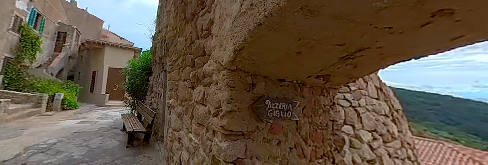 L'immagine mostra una strada stretta e acciottolata a Giglio Castello. Il cartello al centro indica la "Pizzeria Giglio"