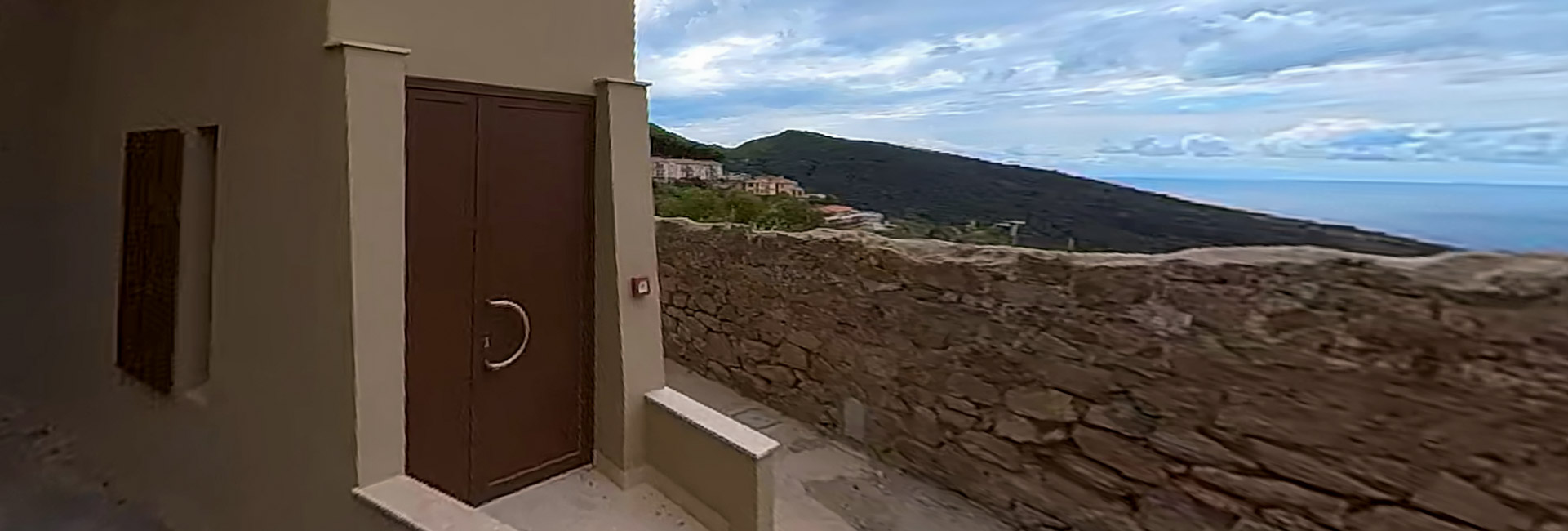 L'immagine ritrae un vicolo con una porta e un muro di pietra, con una vista panoramica sul mare