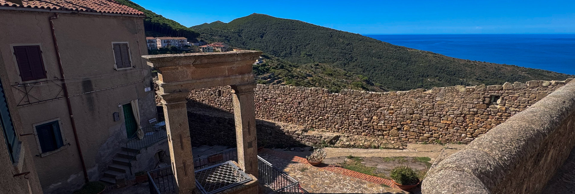 La foto mostra un cortile in pietra di un borgo antico, con un pozzo sormontato da un arco in pietra e due colonne al centro. Sullo sfondo si vedono colline e mare.