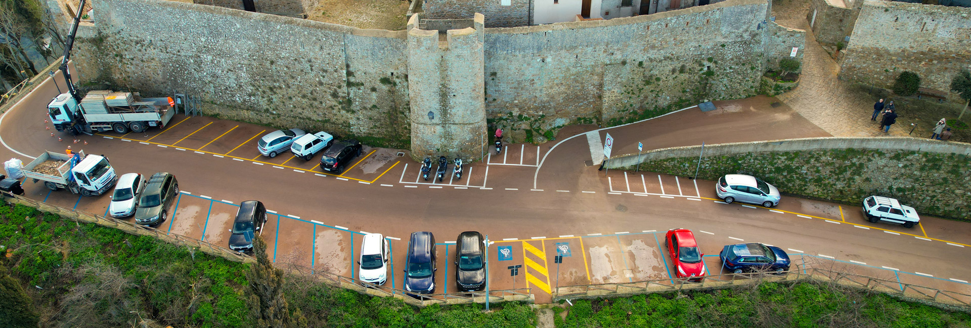 L'immagine mostra una vista aerea di un parcheggio situato accanto a un'antica cinta muraria in pietra