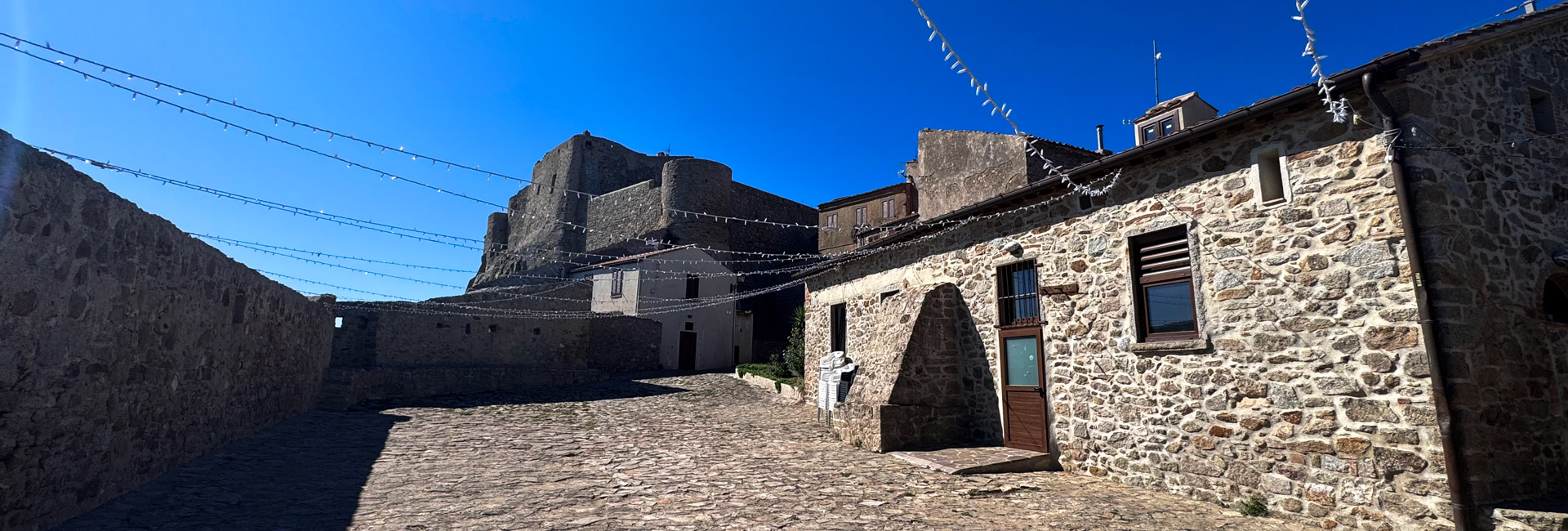 La foto mostra una piazza in pietra nel centro storico del borgo. Sulla sinistra c'è un muro di pietra, mentre sulla desta una casa in pietra.
