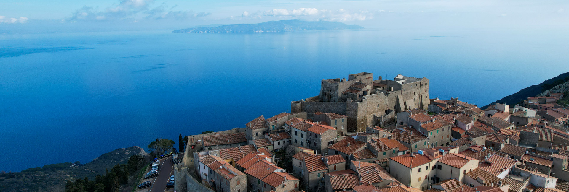 L'immagine mostra la Rocca di Giglio Castello, in una vista aerea che cattura anche una parte del borgo. Sfondo mare ed l'Argentario.